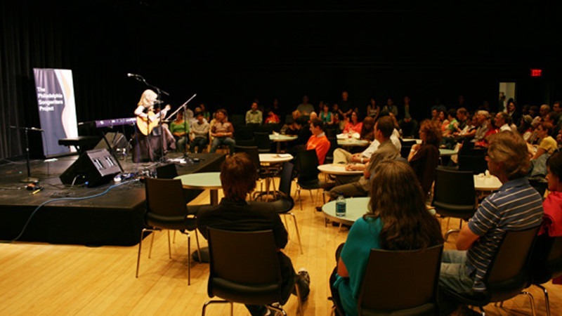 A large group of people listen as a performer plays a song on her guitar.